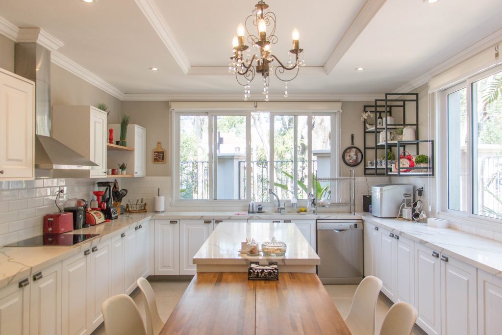 cozinha provençal planejada - beneficios dos armários e a cozinha planejada - cozinha escandinava. A foto mostra uma cozinha planejada em U com ilha no meio, com mesa em madeira maciça e espaço para 8 cadeiras. A cozinha é em tons claros, no formato provençal, com lustre no estilo mais clássico. Os eletros e utensílios ficam a mostra, reforçando o conceito de tudo a mão.